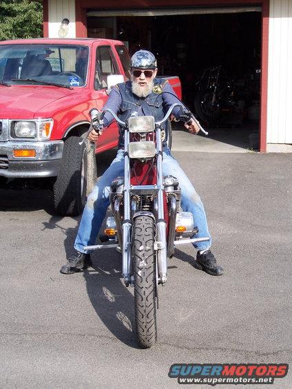 Man with beard and sunglasses sits on a motorcycle in front of a red pickup truck and an open garage