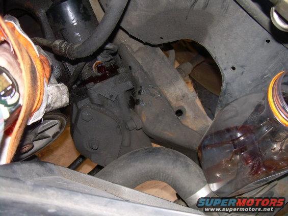 close-up of a leaky power steering pump with red fluid dripping, and a bottle of fluid nearby