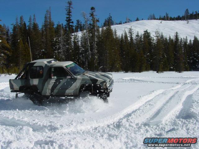 Camouflaged pickup truck driving through deep snow with tire tracks visible