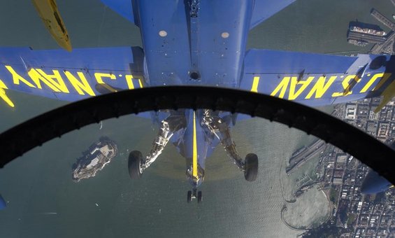 View from cockpit of a U.S. Navy Blue Angels jet flying over water, with a carrier visible below