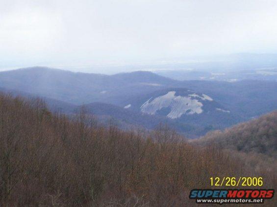 Mountain landscape with a large, light-colored rock formation in the distance, under a hazy sky. Bare trees in the foreground
