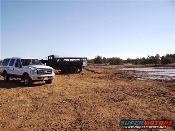 White Ford Excursion parked next to a green military truck in a dirt field with trees in the background