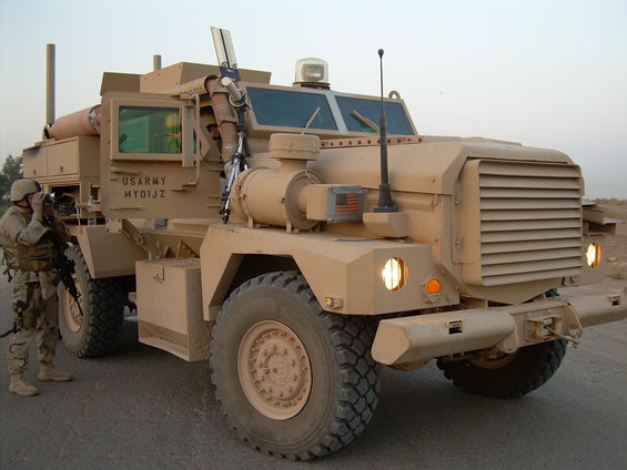 Tan armored military truck with US ARMY MY01JZ markings, front 3/4 view, soldier with rifle standing beside it