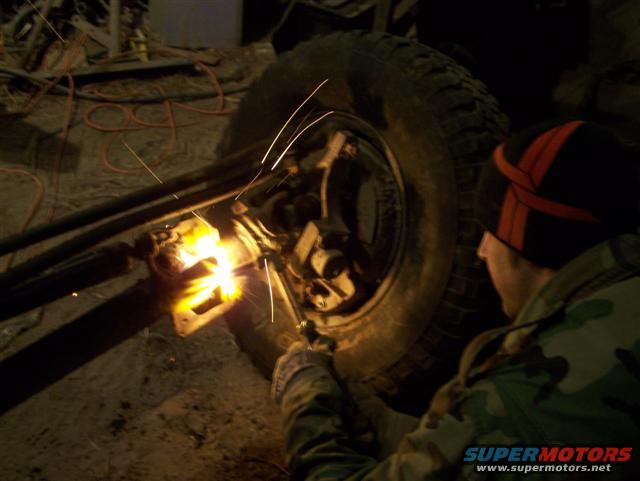 Man in camouflage jacket and beanie welding on a vehicle's axle near a large tire