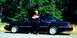 1997 Ford Crown Victoria - A man poses beside a dark-colored sedan parked on the side of a road with a wooded area behind it.