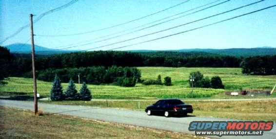 Dark sedan driving on a road with a field and trees in the background