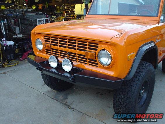 Front view of an orange 1977 Ford Bronco with a black bumper and aftermarket lights. Another vehicle is visible in the background