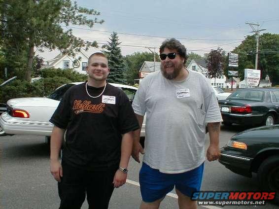 Two men stand in a parking lot with several cars visible in the background