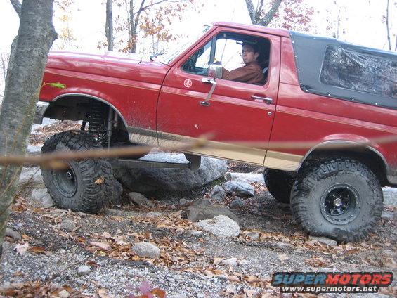 Red 1990 Ford Bronco with a black soft top, lifted suspension, and large off-road tires, navigating rocky terrain
