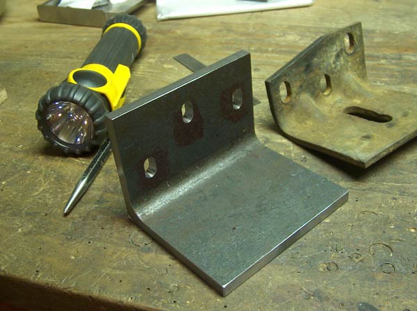 Two metal brackets, one new and one rusty, with a flashlight and scribe on a workbench