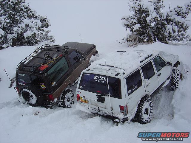 Brown SUV and white Jeep Cherokee stuck in deep snow, trees covered in snow in background