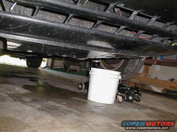 Underneath a car, a white bucket and a jack are positioned near the rear tire. A green hose is visible on the ground