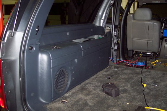Interior view of a vehicle showing a speaker enclosure on the left, exposed wiring, and a removed seat on the right