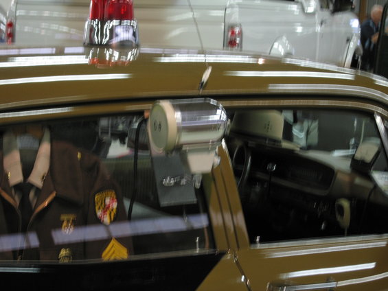 Interior of a police car showing a uniform on the passenger seat, a spotlight mounted on the window, and a red light bar on the roof