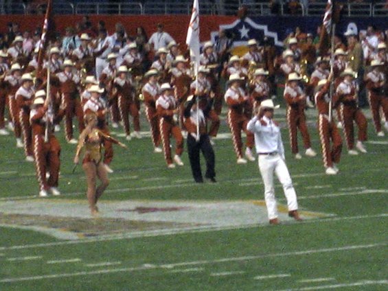 Marching band members in maroon and white uniforms and cowboy hats, with a dancer in gold, on a football field