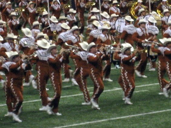 Marching band members in cowboy hats and brown uniforms play instruments on a green field