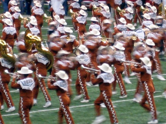 Marching band members in cowboy hats and brown uniforms play instruments on a football field