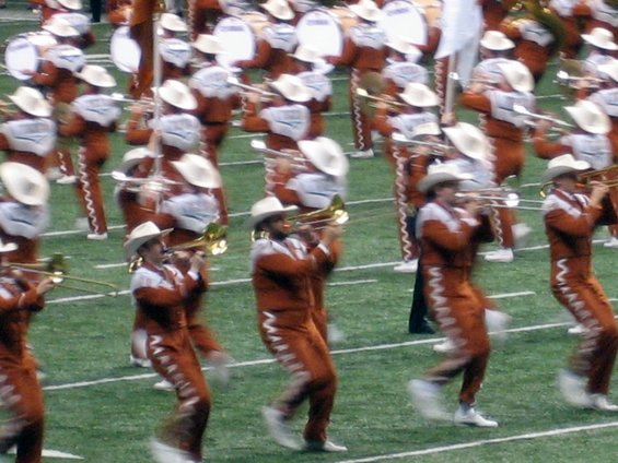 Marching band members in orange uniforms and cowboy hats play instruments on a football field