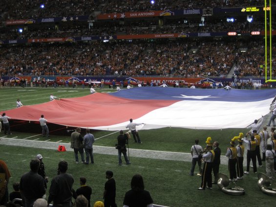 Texas flag being held by people on a football field with a marching band and spectators in the background