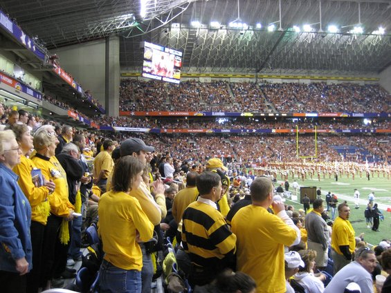 People in yellow shirts watch a football game in a stadium, with a marching band on the field