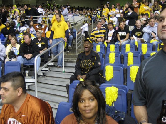 Spectators in blue stadium seats with yellow pom-poms, some wearing team colors