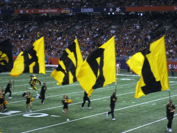 Football field with cheerleaders and large yellow and black flags, San Antonio Alamo Bowl banner in background