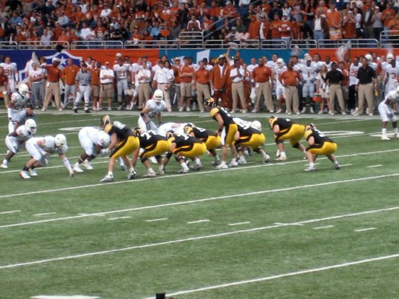 American football game with players in white and yellow uniforms on the field, spectators in the stands
