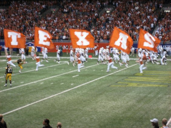 Football players run onto a field, holding large orange flags spelling 'TEXAS'. Spectators fill the stands