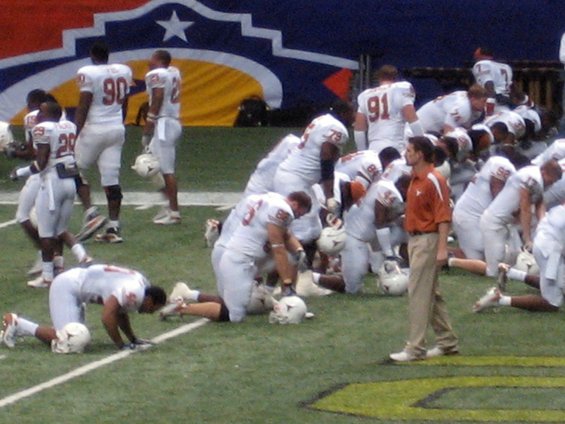 Football players in white uniforms kneeling on a field with a coach watching