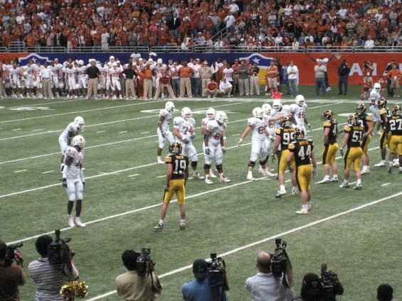 Football players in white and yellow uniforms on a field, with spectators in the background