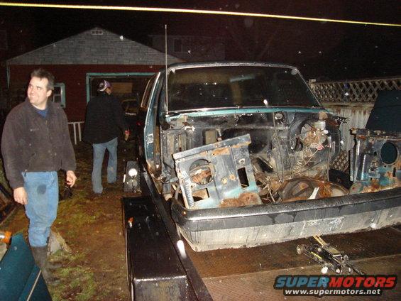 car body stripped down on a trailer at night, two people standing nearby