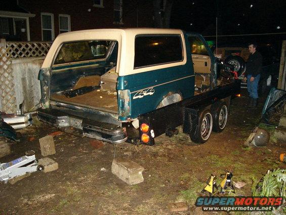 Rear view of a teal and white Ford Bronco that has been converted to a flatbed truck. It's parked outside at night