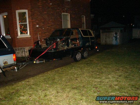 Dark green Ford Bronco body on a black trailer, front end removed, red straps securing it