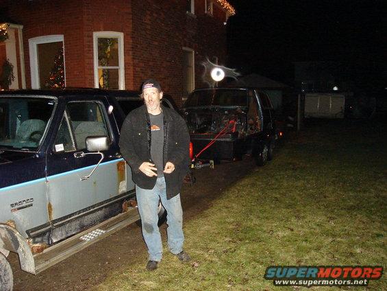 Man standing next to a rusty 1990 Ford Bronco, with another vehicle on a trailer behind it