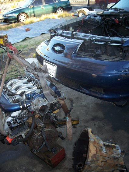 Engine removed from dark blue car, transmission nearby. Green car in background
