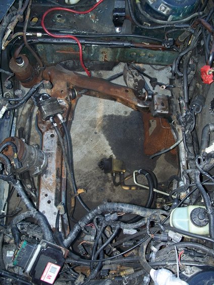 Engine bay with exposed wiring, hoses, and rusty suspension components. A brake fluid reservoir is visible