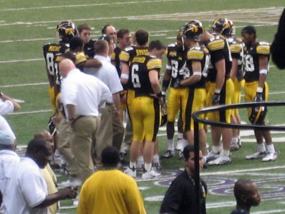 Football players in black and yellow uniforms huddle with coaches on a green field