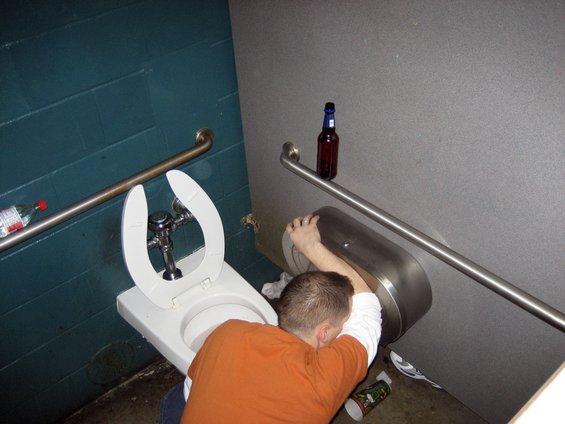Man passed out in a bathroom stall next to a toilet with a beer bottle on the wall