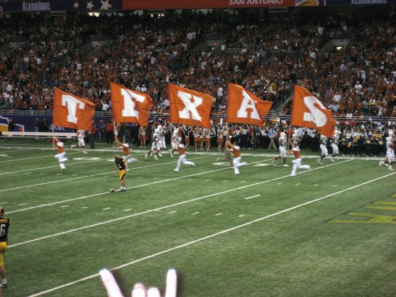 Football field with orange flags spelling TEXAS, players running, and a crowd in the stands