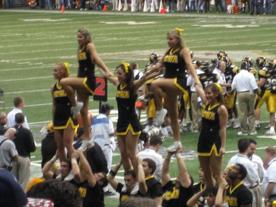 Cheerleaders in black and yellow uniforms form a pyramid on a football field during a game