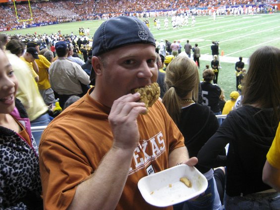 guy eating fried chicken in stadium seats, people around him, football field in background