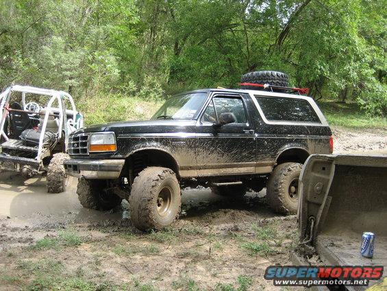 Muddy 1996 Ford Bronco with spare tire on roof rack, parked next to an off-road buggy in a muddy area