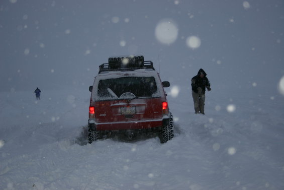 Red Jeep SUV with roof rack and spare tire stuck in deep snow, rear view