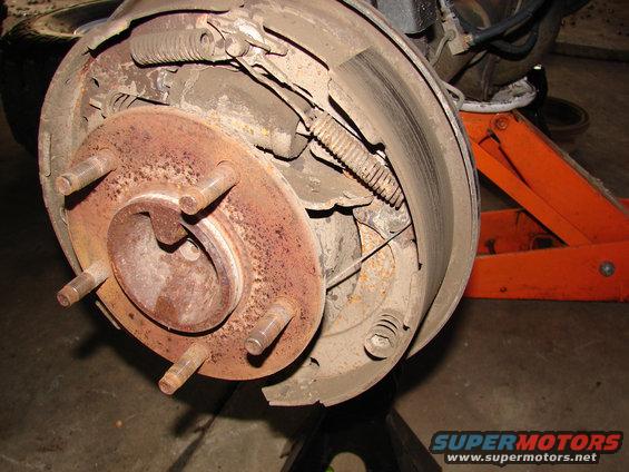 Close-up of a rusty rear drum brake assembly with brake shoes and springs visible. A hydraulic jack is in the background
