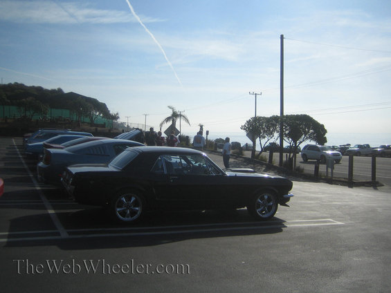 Black 1965 Ford Mustang parked in a lot with other cars, ocean in background