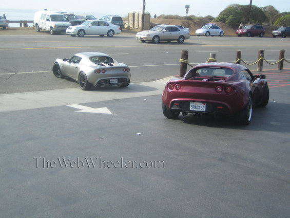 Two Lotus Elise sports cars parked near a road with the ocean in the background. One is silver, the other is red