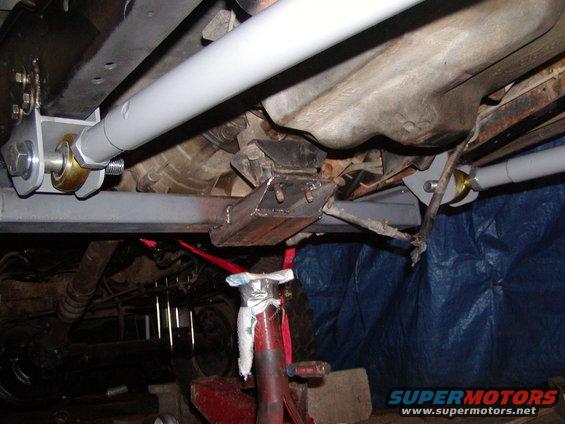 Underbody view of a vehicle showing modified suspension components, including grey painted bars and brackets, with a red jack stand visible