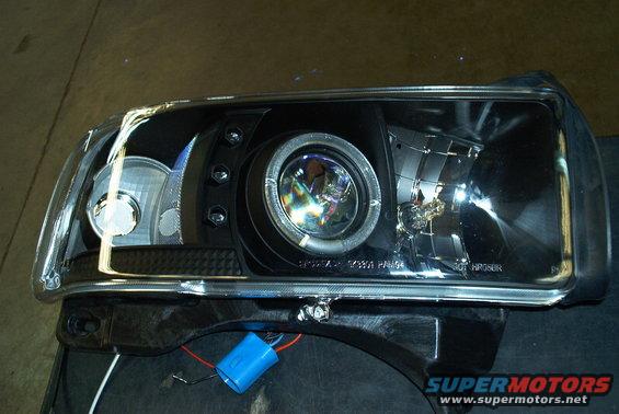 Black headlight assembly with projector lens and halo ring, showing wiring and a blue capacitor
