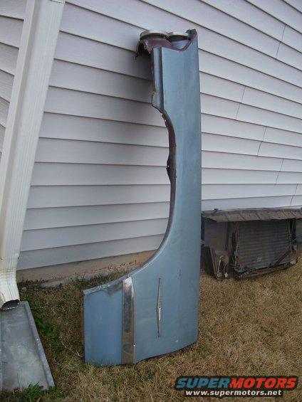 Light blue car fender with chrome trim and a rusty radiator unit on the grass next to a house