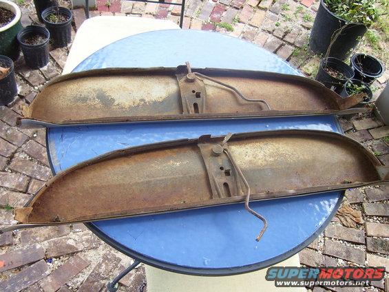 Two rusty, curved metal car parts with mounting brackets and wires on a blue table